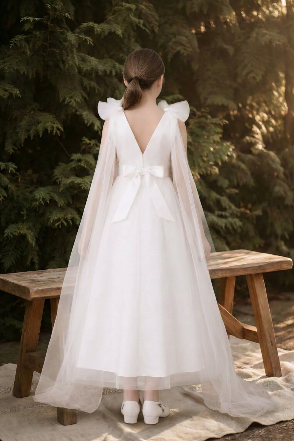 Flower girl standing in a forest environment, displaying the beautiful drape of the long tulle cape on her ivory white dress.