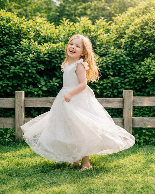A smiling little flower girl twirling outdoors in an ivory white dress with a sparkly tulle skirt and flutter sleeves.