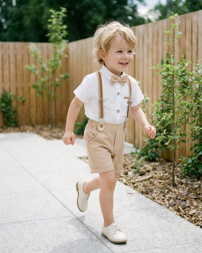 A happy little ring bearer walking outdoors next to a wooden fence wearing a smart khaki dress shorts set with suspenders