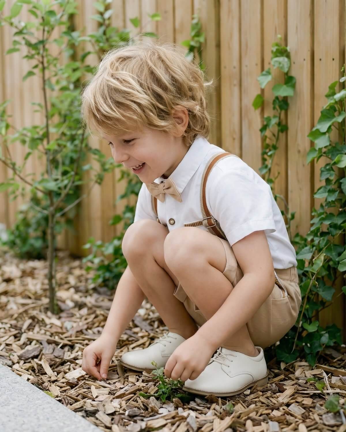 Playful candid shot of a toddler boy squatting outdoors in a breathable short-sleeve cotton shirt and beige suspender shorts combo