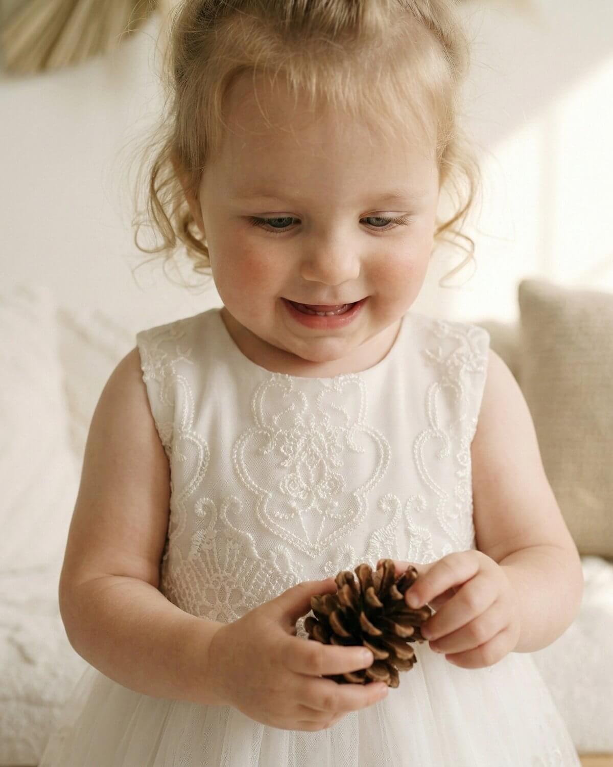 Close-up of baby girl holding a pinecone, showing the delicate embroidered lace bodice details of the ivory Seraphina dress
