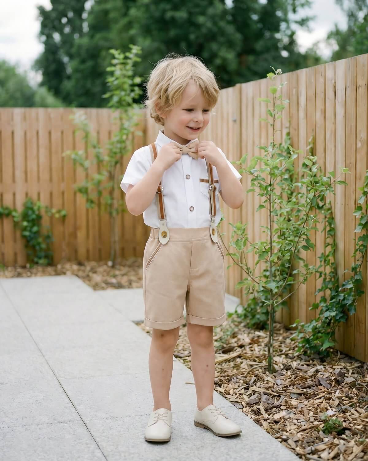 Full length view of a small boy standing outdoors in a boho style khaki suspender shorts attire, adjusting his matching beige bowtie