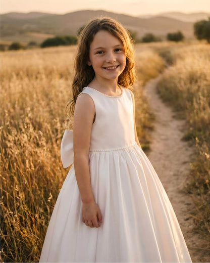 Side profile view of a girl smiling in The Grace ivory satin wedding flower girl gown in a sunny field