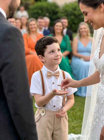 Cute ring bearer wearing beige pants, brown suspenders, and a bow tie handing rings to the bride at an outdoor wedding ceremony
