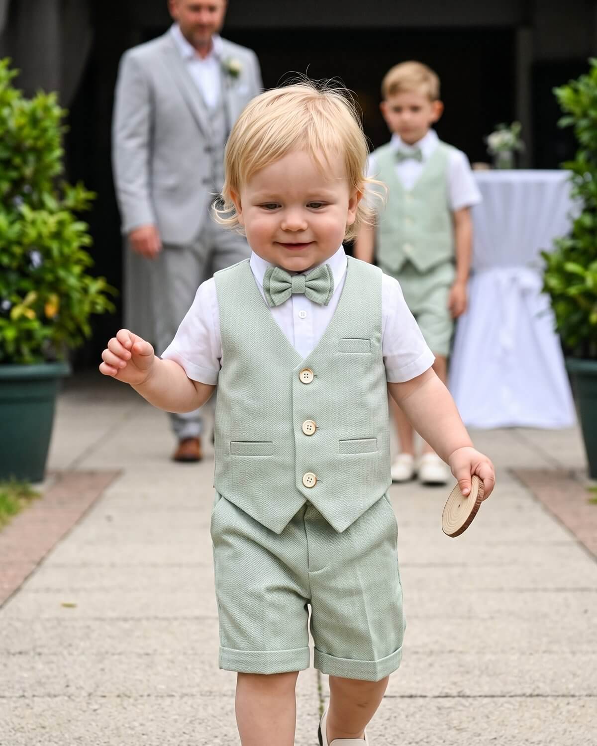 Cute toddler ring bearer wearing a sage green vest and shorts suit set at an outdoor wedding.