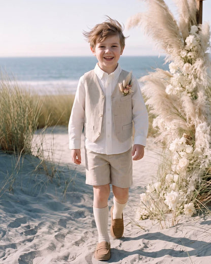 Young boy smiling on a sandy beach wearing an oatmeal 3-piece linen-lyocell vest, white shirt, and shorts set by a pampas grass arch.