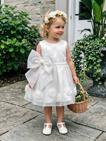 A cute toddler flower girl wearing an ivory white satin dress with a large side bow, a pearl beaded waistband, and a floral crown, holding a small woven basket in a garden.