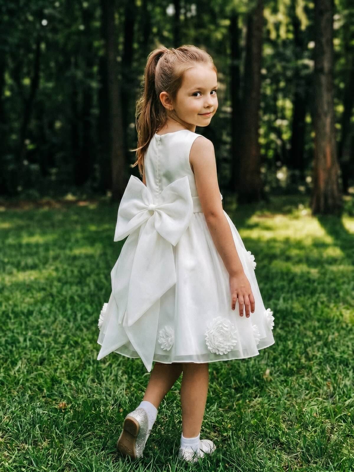 Young flower girl wearing a beautiful white dress featuring a large back bow and 3D floral appliques on the skirt, standing in a garden.