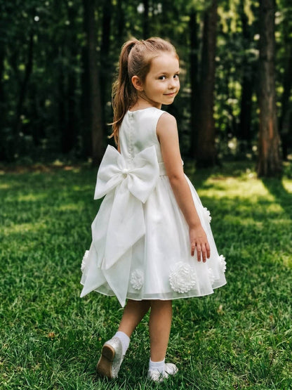 Young flower girl wearing a beautiful white dress featuring a large back bow and 3D floral appliques on the skirt, standing in a garden.