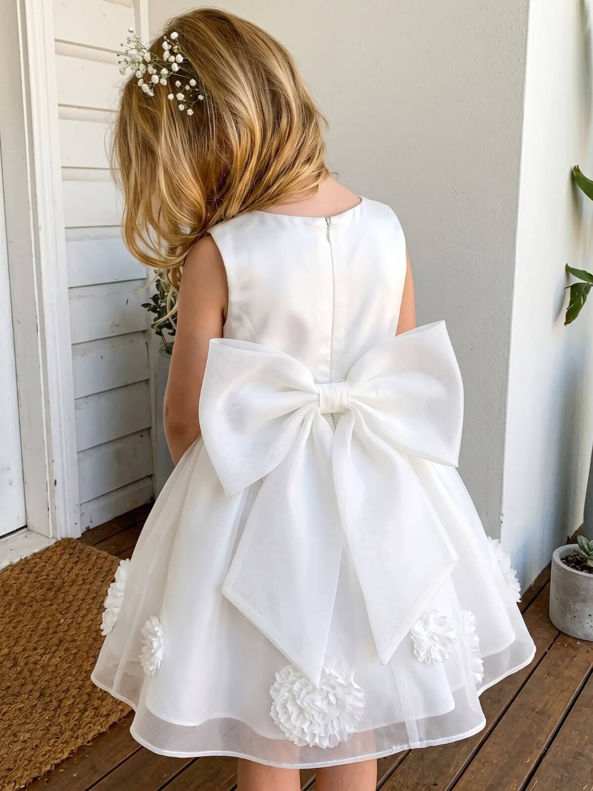 Back view of a young blonde flower girl with a baby's breath hairpiece, wearing a luxury white satin dress featuring an oversized statement back bow and 3D floral hem.