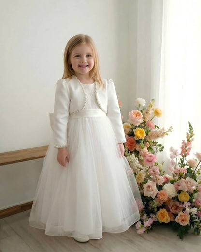 Full-length portrait of a smiling little girl wearing an ivory satin pearl-trimmed shrug cover-up paired with a floor-length tulle formal wedding dress in a floral studio.