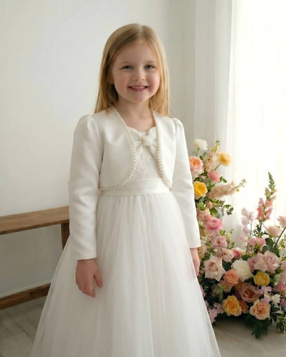 Front view of a young flower girl standing indoors, wearing a premium ivory wedding satin cropped bolero jacket with elegant pearl beaded trim over a white lace communion dress.