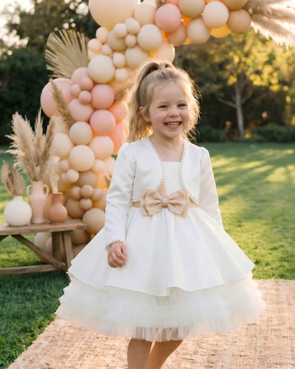 A little girl laughing outdoors in front of a balloon arch, styled in an ivory satin formal shrug with pearl collar over a birthday party dress with a tan bow.