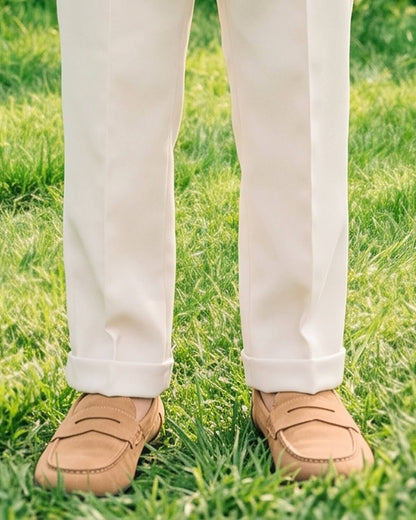 Detail shot of Little Ivory boy's formal cream beige dress pants hem. Shows the neatly cuffed trouser hem with a sharp crease, paired with tan suede loafers on green grass for an outdoor wedding.