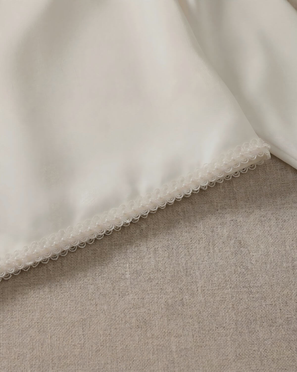 Macro close-up shot of the exquisite hand-crafted pearl beaded scalloped lace trim lining the bottom edge of the smooth ivory satin jacket.