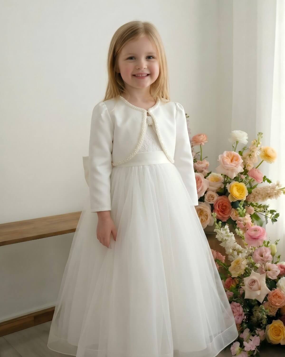 Three-quarter portrait of a little girl wearing a white formal satin shrug cover-up with pearl border decoration next to a pastel floral backdrop.